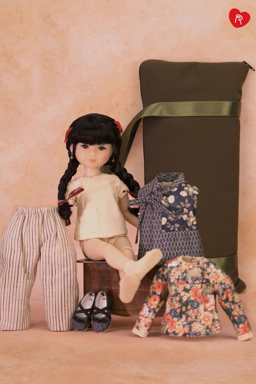 A doll with black braided hair sits on a small wooden stool in a styled studio setup, surrounded by clothing and accessories including striped pants, black shoes, a floral jacket, and a large olive green bag against a warm beige backdrop.