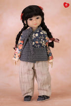 A doll with long black braided hair and red hair ties, wearing a dark floral vest over a gray patterned top, striped pants, and dark shoes, posed against a soft peach studio background.