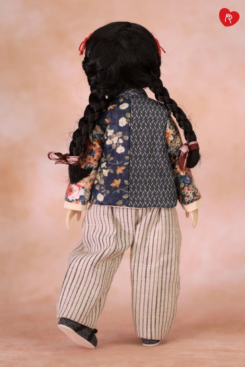 A doll viewed from the back wearing a dark navy patterned vest over a floral long-sleeve top, cream and gray vertically striped pants, and black shoes, against a soft peach studio background.