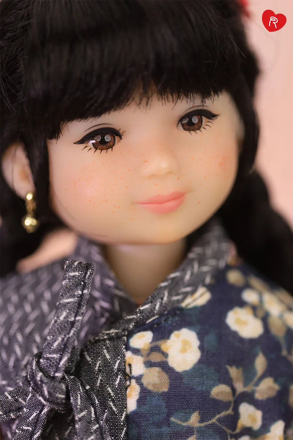 Close-up of a doll with dark hair, large brown eyes, and a floral dark blue top with white flowers, wearing a small gold earring.