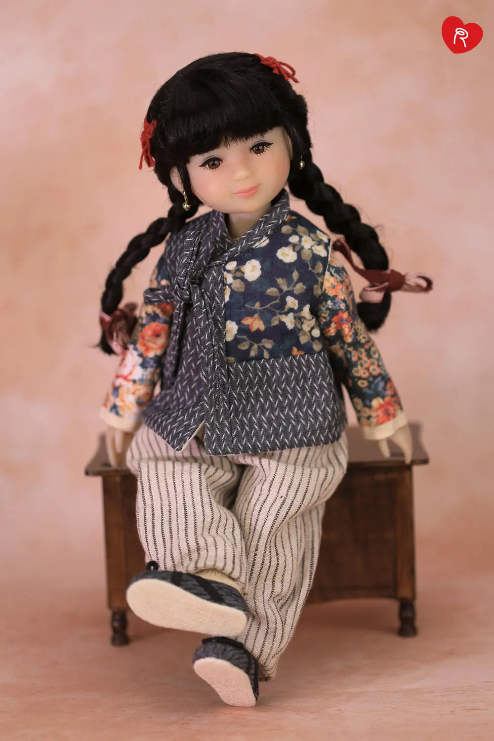 A doll with long black braided hair sits on a small wooden stool against a soft pink studio background. The doll is wearing a dark floral jacket, striped pants, and dark shoes.