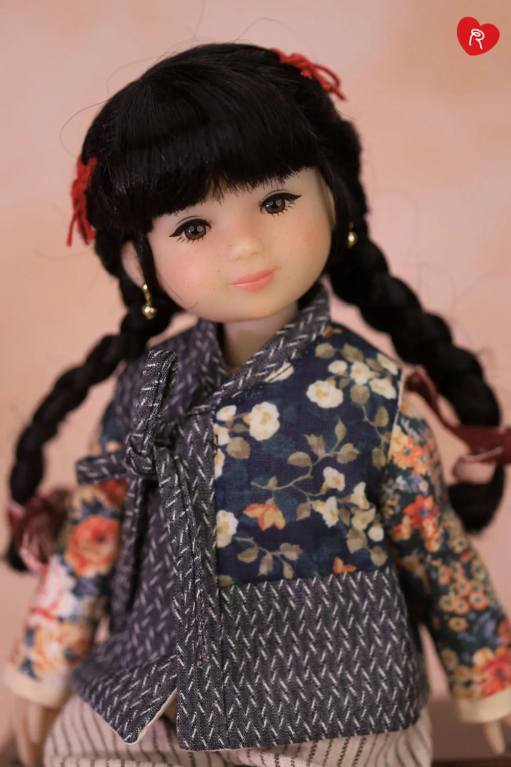 A close-up of a doll with long black braided hair and a floral embroidered jacket, set against a soft pink background.