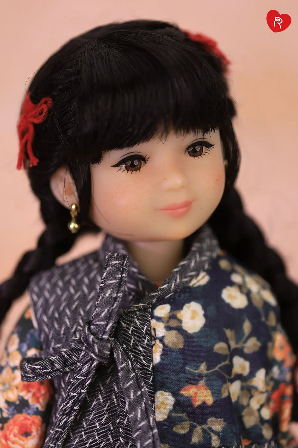 A close-up portrait of a doll with dark hair in braids, wearing a navy floral top and a gray patterned bow tie, set against a soft pink background.