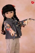 A doll with long black braided hair and a red hair tie, wearing a dark patterned vest over a floral long-sleeve top and striped pants, posed against a soft peach-pink background.