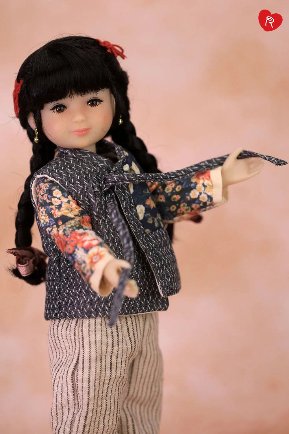 A doll with long black braided hair and a red hair tie, wearing a dark patterned vest over a floral long-sleeve top and striped pants, posed against a soft peach-pink background.