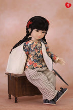 A doll with black braided hair and red hair bows sits on a small wooden box, wearing a floral long-sleeve top, striped pants, and dark shoes against a soft peach background.