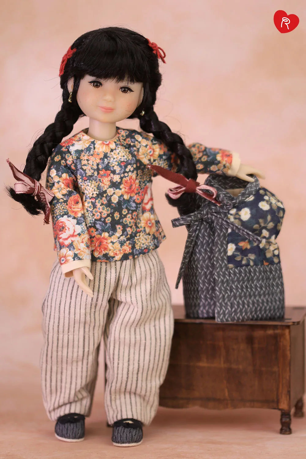 A doll with long black braids and a red hair bow stands next to a small wooden table, wearing a floral long-sleeve top and striped pants, with a matching floral bag beside it.