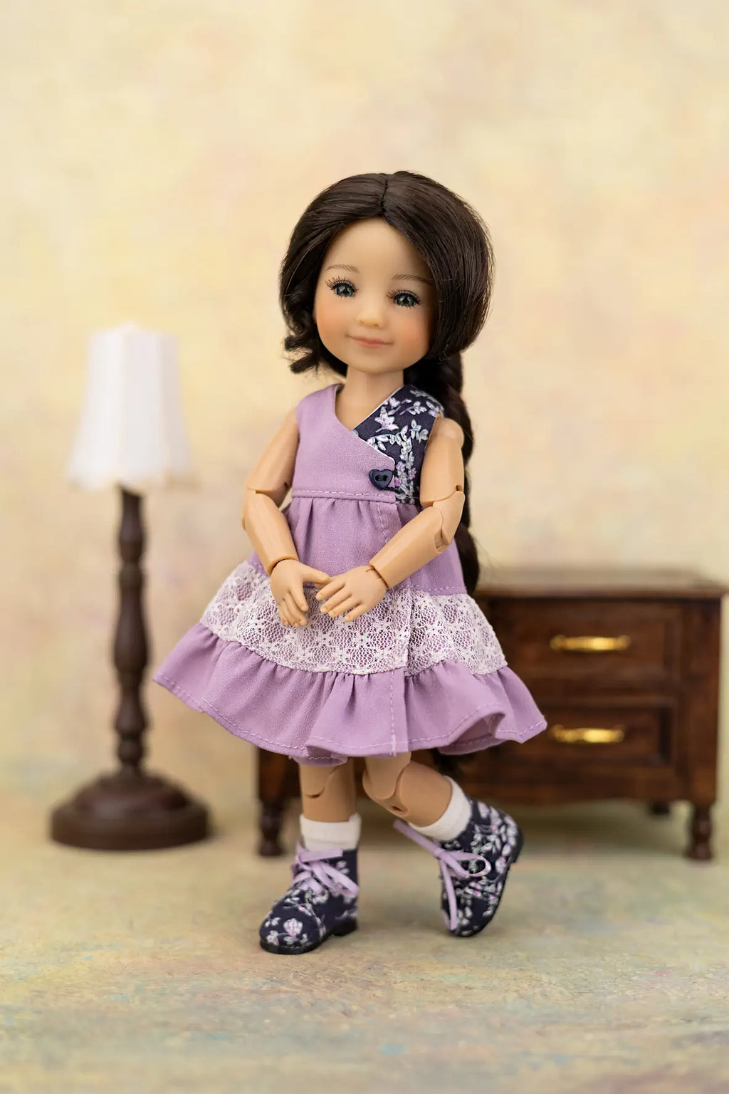 A doll with dark brown hair wearing a lavender sleeveless dress with a white lace hem, patterned shoes, and white socks, posed in front of a miniature lamp and cabinet backdrop.