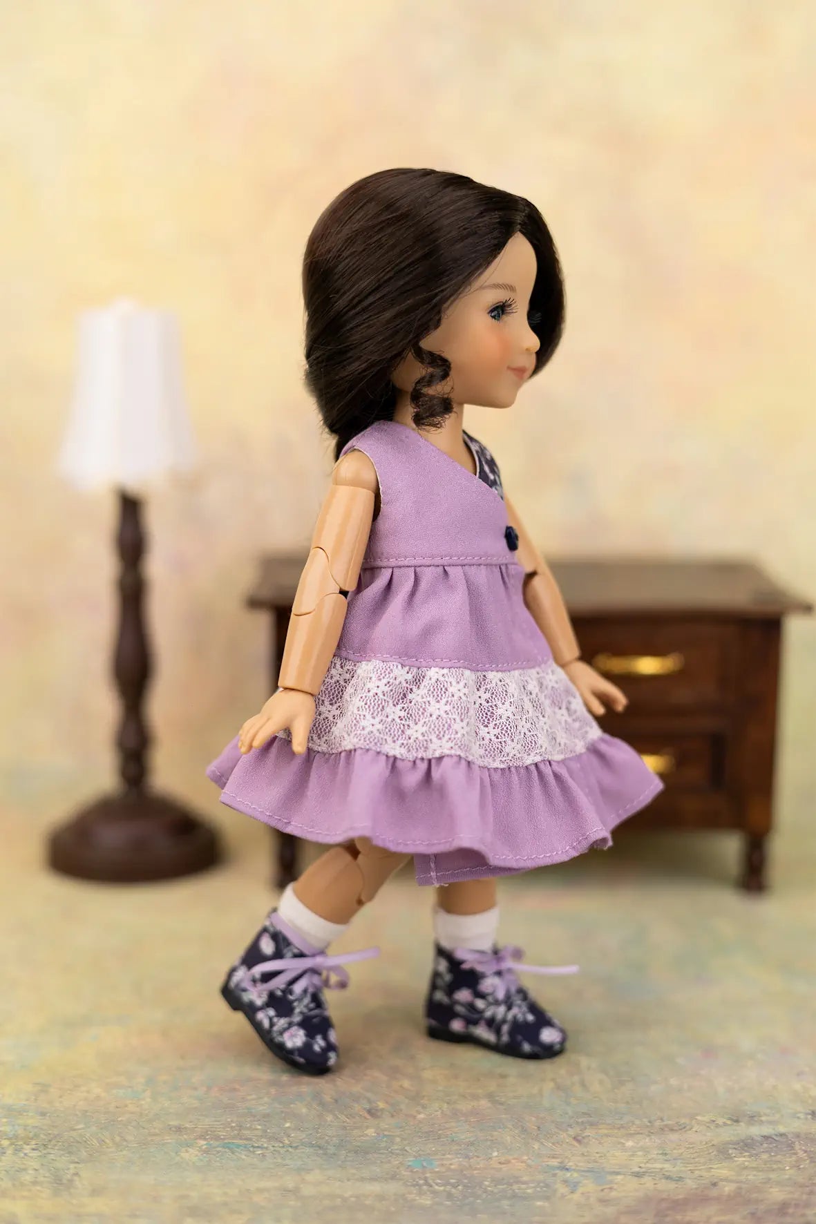 A doll with dark brown hair wearing a lavender sleeveless dress with a white lace ruffle, white socks, and dark floral shoes, posed in a miniature room setting with a small lamp and cabinet in the background.