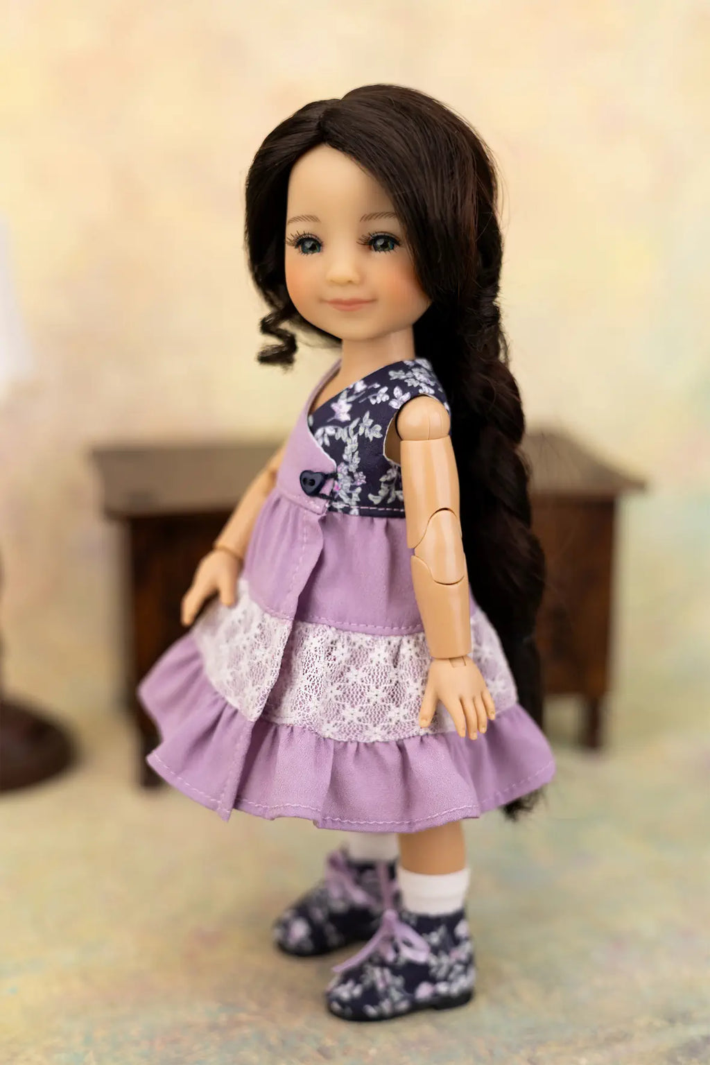 A doll with long dark brown hair wearing a lavender and navy floral dress with white lace trim and matching floral socks and shoes, posed against a softly blurred indoor background.