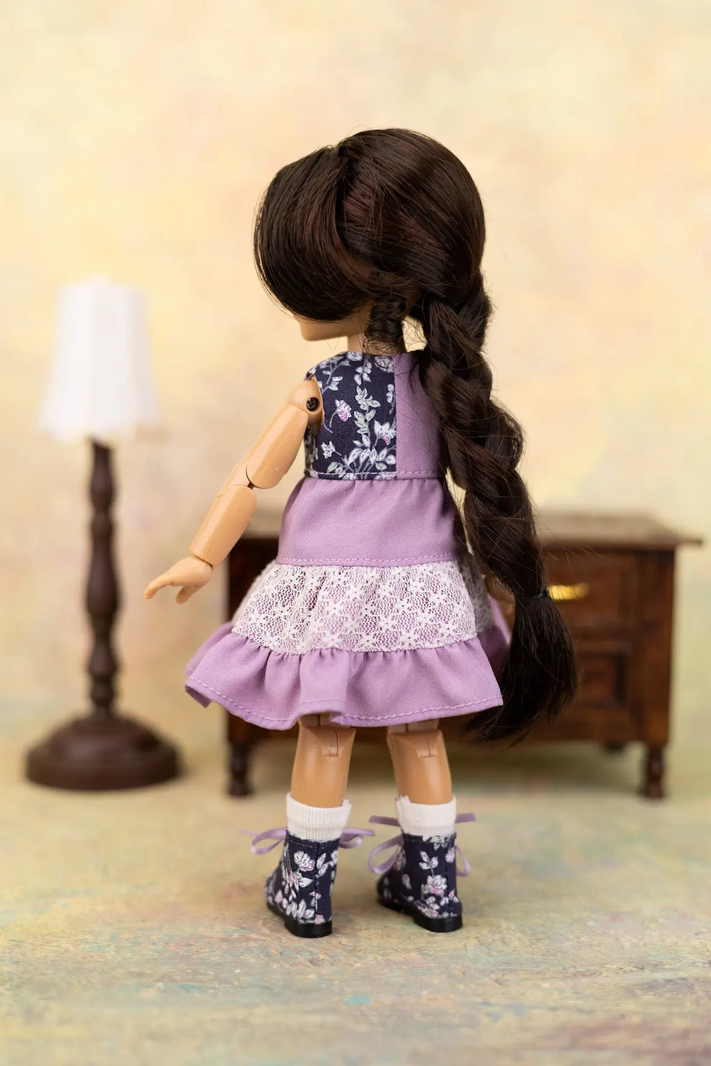 A doll viewed from the back wearing a lavender and navy floral dress with a white lace ruffle, white socks, and dark floral boots, standing in a miniature room setting with a lamp and cabinet.