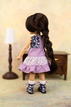 A doll viewed from the back wearing a lavender and navy floral dress with a white lace ruffle, white socks, and dark floral boots, standing in a miniature room setting with a lamp and cabinet.