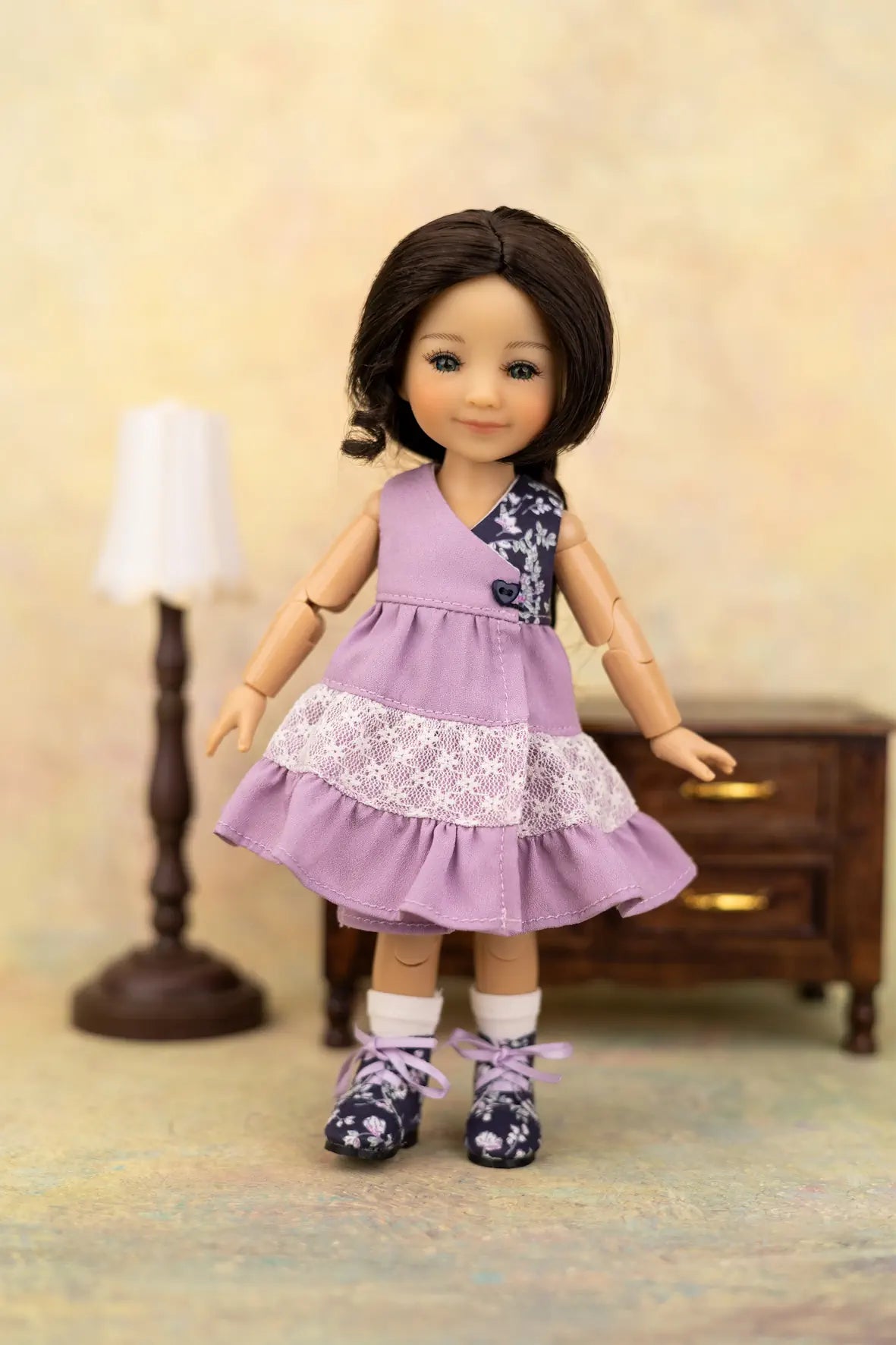 A doll with dark brown hair wearing a lavender sleeveless dress with white lace trim, white socks, and dark floral shoes, posed in front of a miniature lamp and cabinet backdrop.