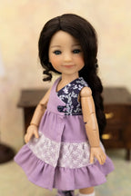 A doll with long dark brown hair wearing a lavender sleeveless dress with a navy floral yoke and a white lace ruffle, posed against a softly blurred indoor background.