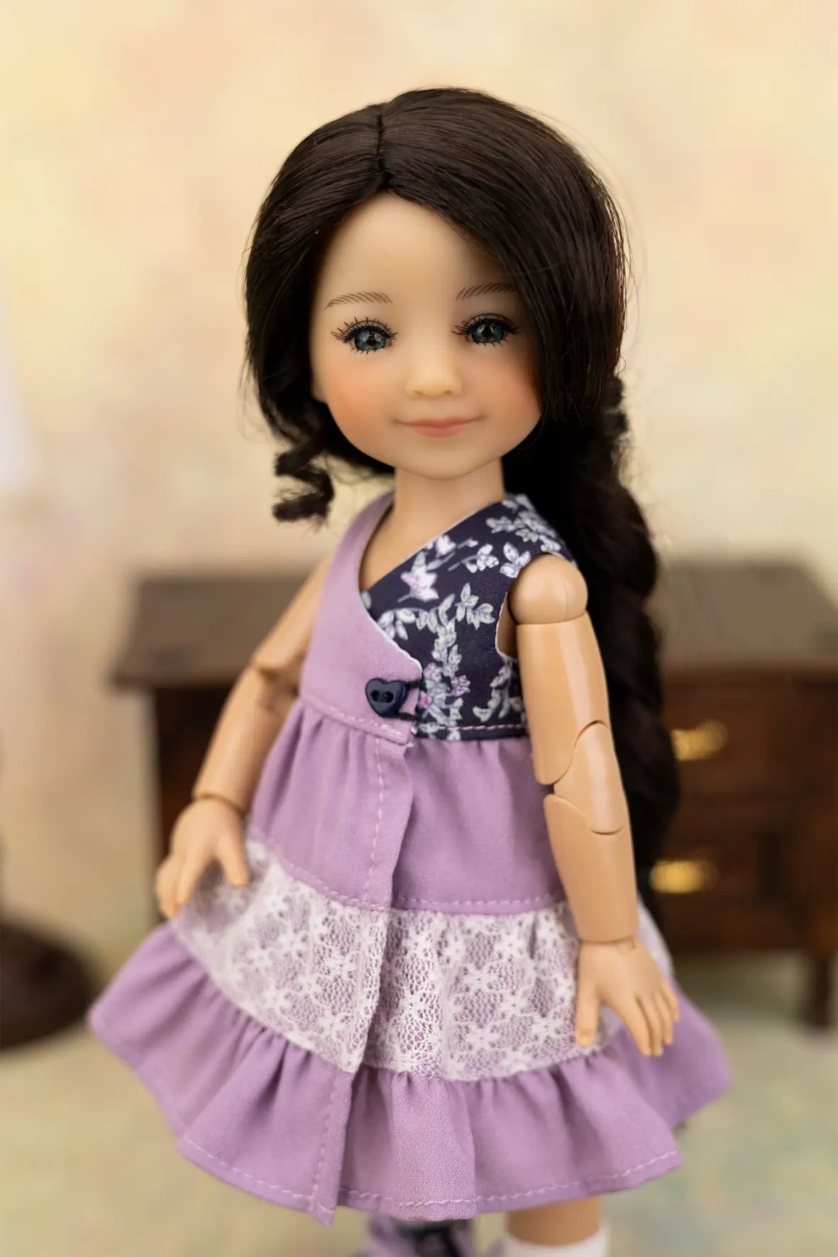 A doll with long dark brown hair wearing a lavender sleeveless dress with a navy floral yoke and a white lace ruffle, posed against a softly blurred indoor background.