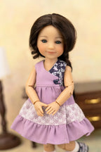 A doll with dark brown hair wearing a lavender sleeveless dress with a floral patterned yoke and a white lace hem, posed against a softly blurred indoor background.