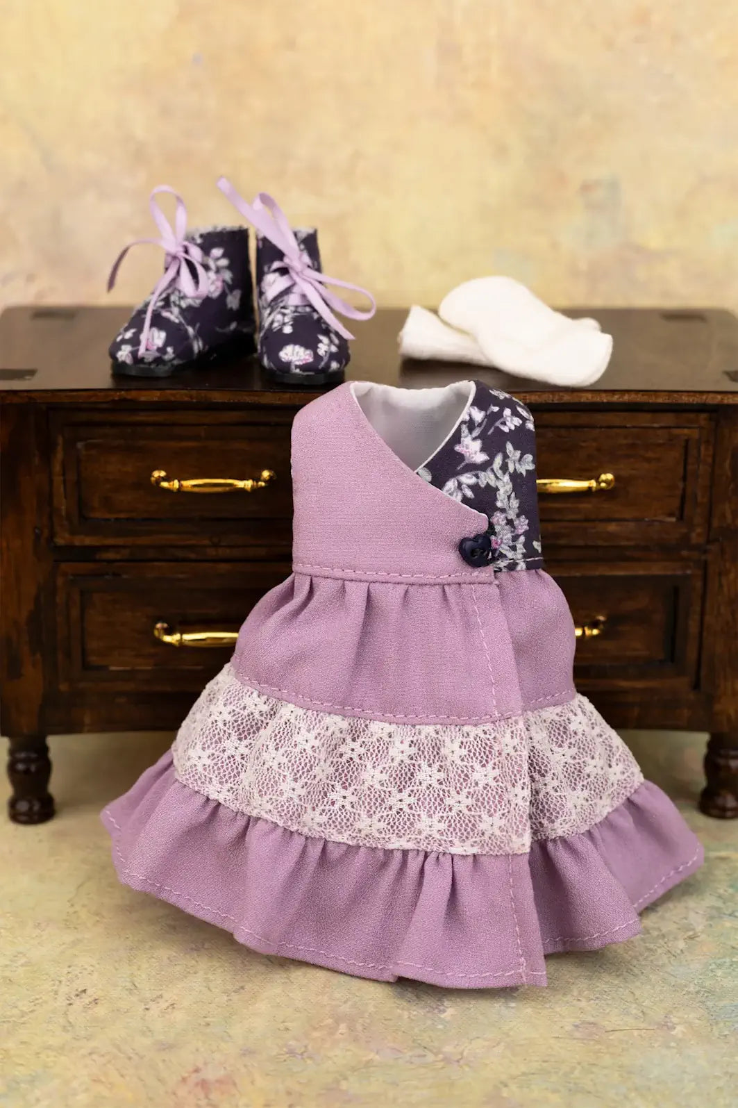 A doll dress in dusty lavender with a white lace ruffle and a floral patterned collar, displayed on a miniature wooden chest with matching floral doll boots and white socks.