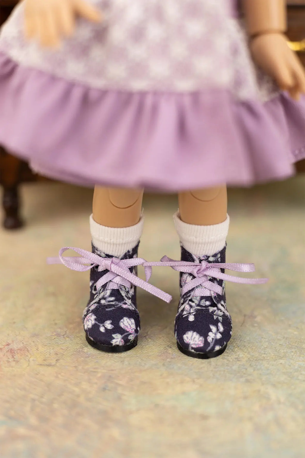 A close-up of a doll's feet wearing dark navy floral lace-up shoes with white socks and a lavender skirt.