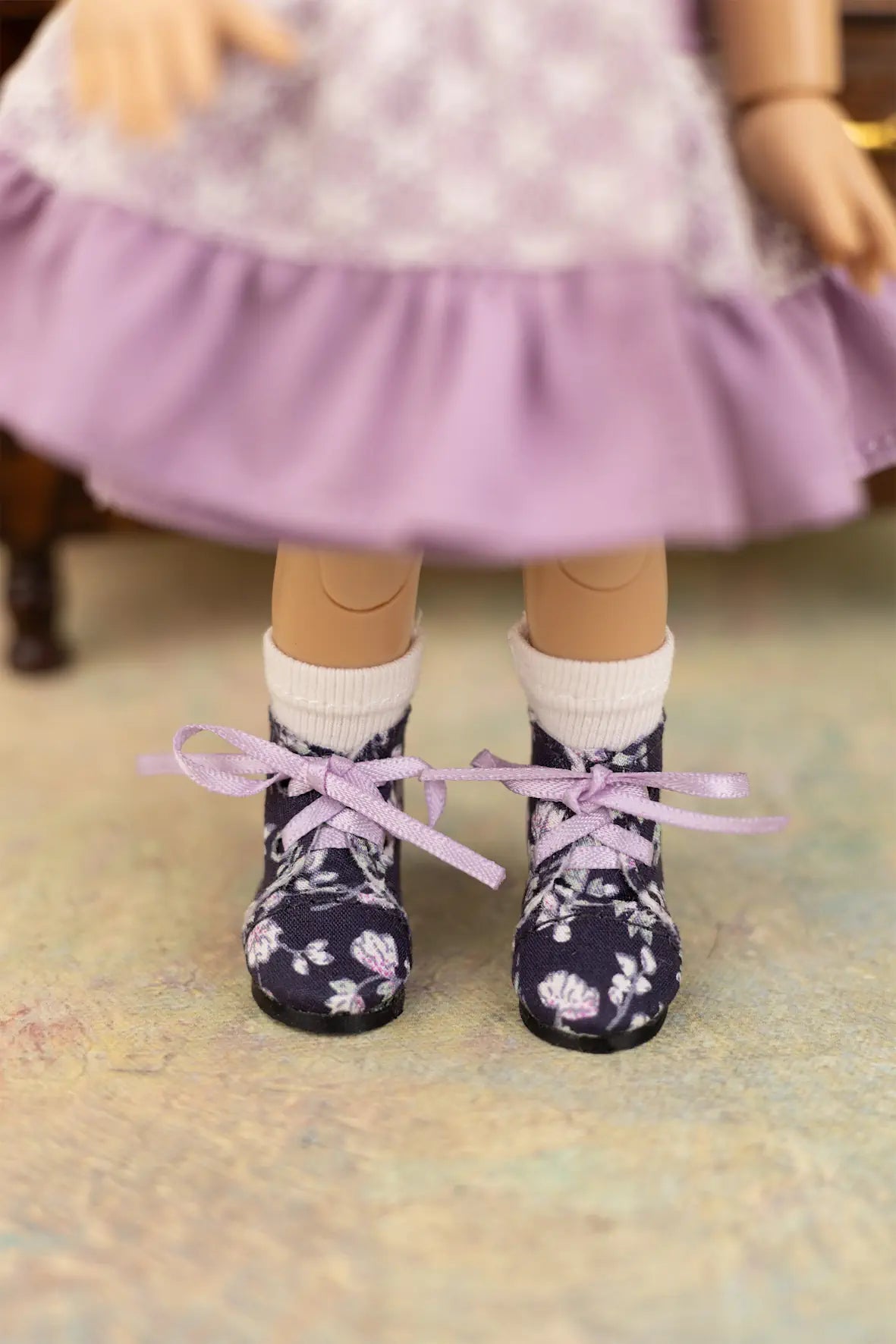 A close-up of a doll's feet wearing dark navy floral lace-up shoes with white socks and a lavender skirt.