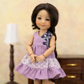 A doll with dark brown hair wearing a lavender sleeveless dress with a floral yoke and white lace trim, posed against a softly blurred indoor background with a lamp and wooden furniture.