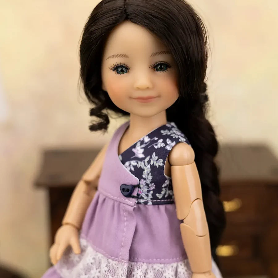 A close-up of a doll with long dark brown hair wearing a lavender sleeveless dress with a floral-patterned yoke and white lace trim, set against a softly blurred indoor background.