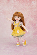 A doll with long auburn hair wearing a yellow floral dress with a white flower applique, pink socks, and yellow sneakers, posed against a pink textured backdrop.