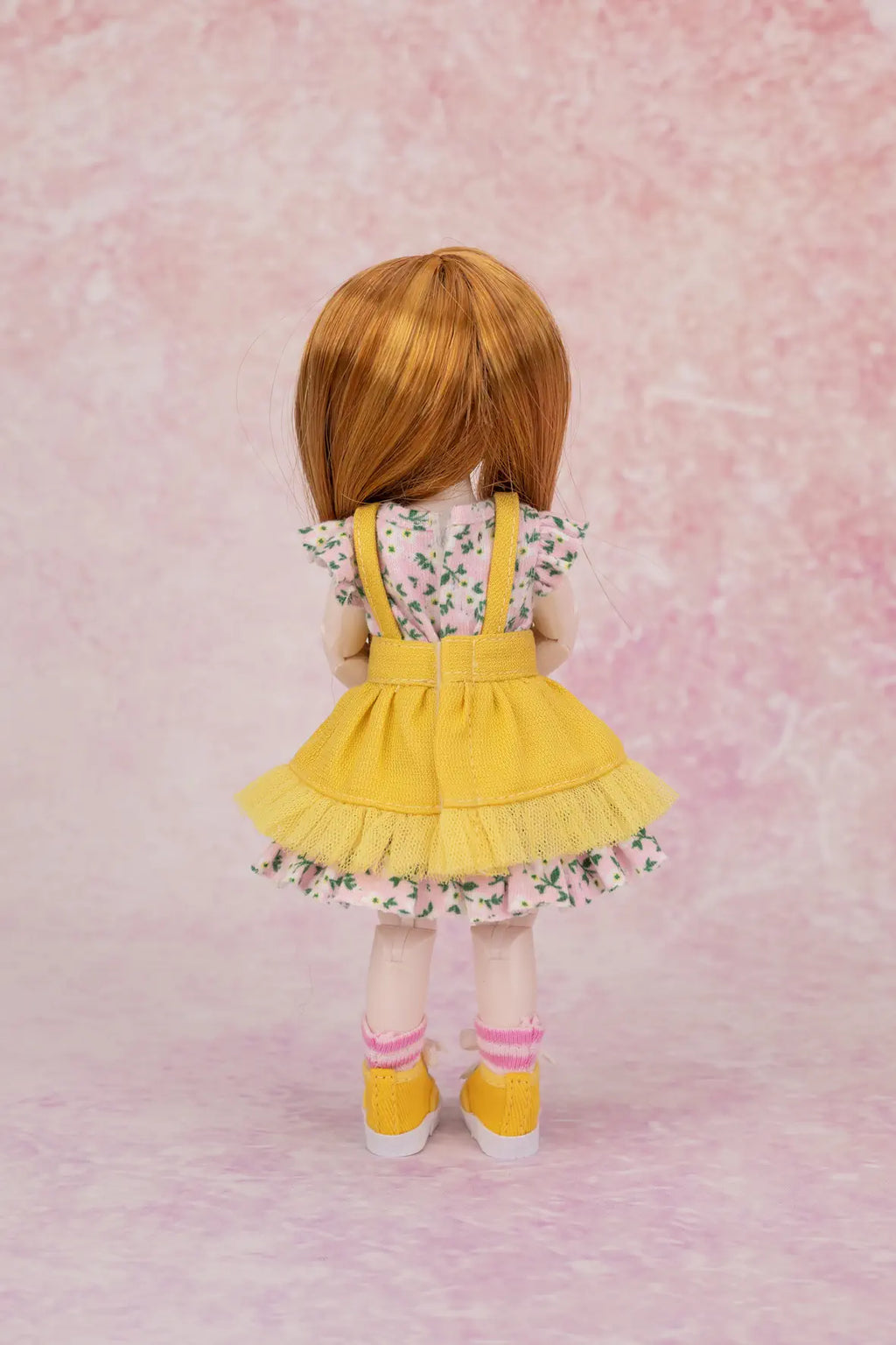 A doll viewed from the back wearing a yellow denim-look pinafore dress over a white floral short-sleeve top, with pink striped socks and yellow shoes, standing against a pink textured backdrop.