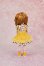 A doll viewed from the back wearing a yellow denim-look pinafore dress over a white floral short-sleeve top, with pink striped socks and yellow shoes, standing against a pink textured backdrop.
