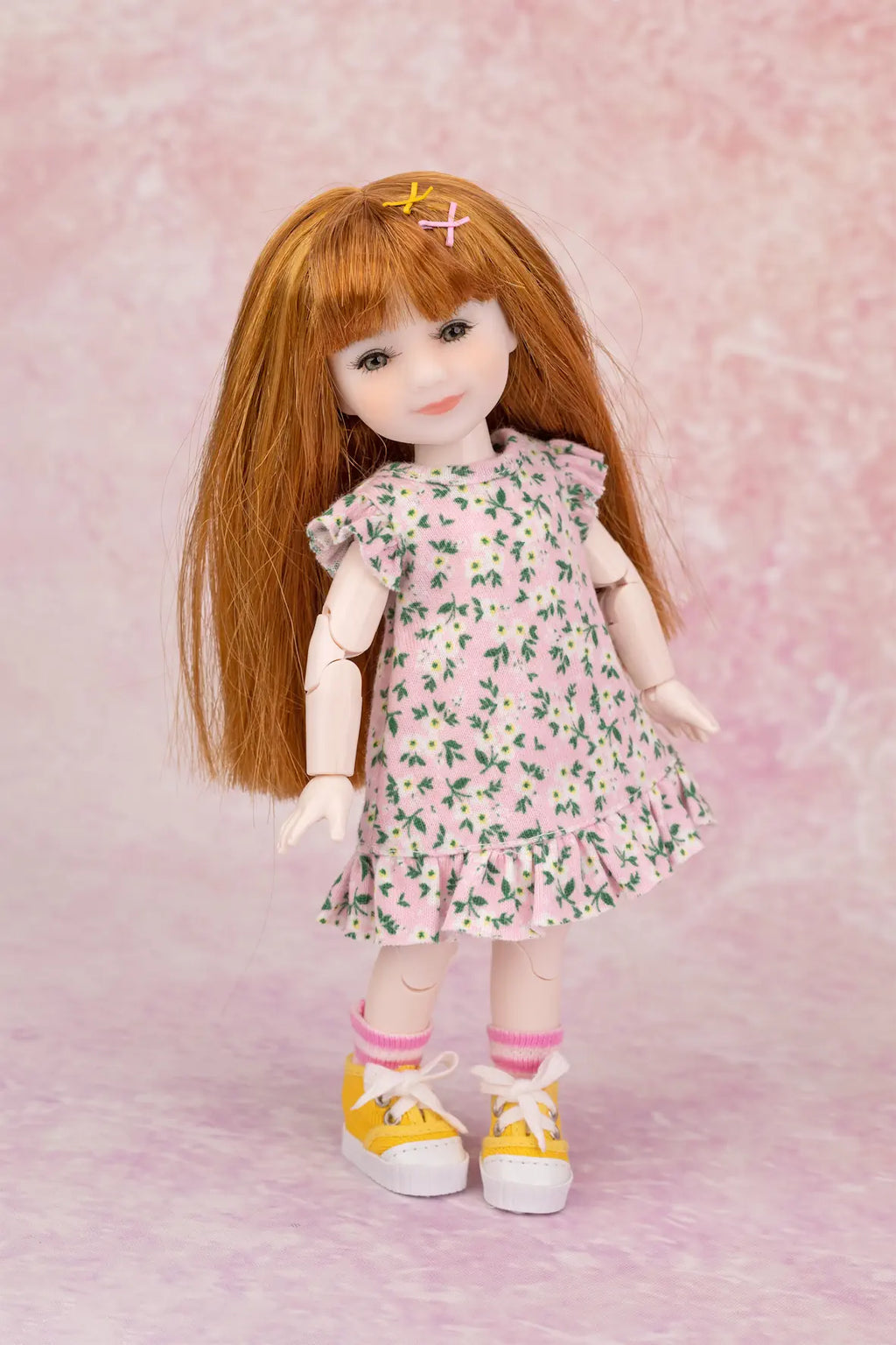 A doll with long auburn hair wearing a pink floral dress, pink socks, and yellow sneakers, posed against a pink textured backdrop.