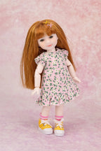 A doll with long auburn hair wearing a pink floral dress, pink socks, and yellow sneakers, posed against a pink textured backdrop.