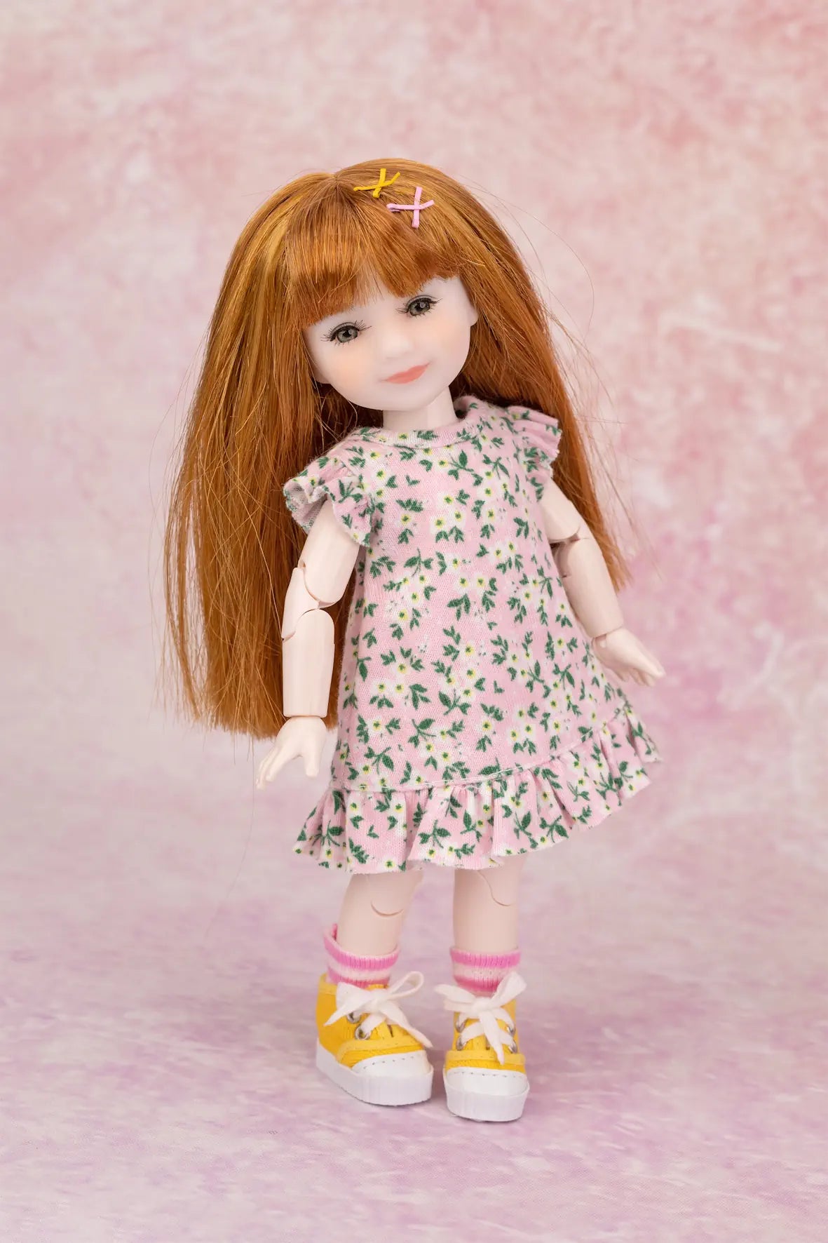 A doll with long auburn hair wearing a pink floral dress, pink socks, and yellow sneakers, posed against a pink textured backdrop.