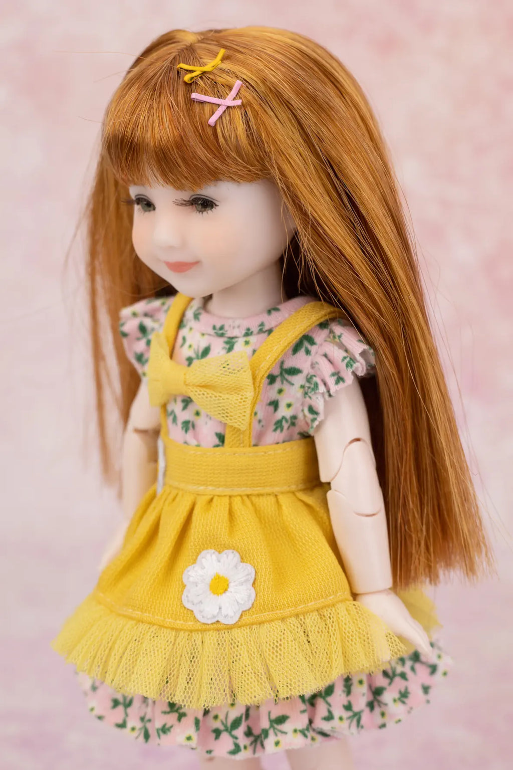 A doll with long auburn hair wearing a yellow pinafore-style dress over a pink floral top, posed against a soft pink background.
