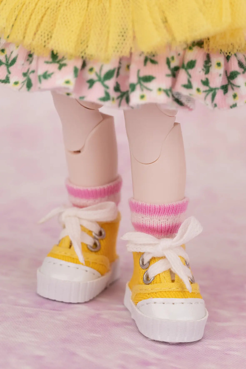 Yellow canvas-style miniature sneakers with white laces and white soles, worn with pink striped socks and a floral skirt, shown on a doll's legs against a pink background.
