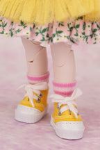 Yellow canvas-style miniature sneakers with white laces and white soles, worn with pink striped socks and a floral skirt, shown on a doll's legs against a pink background.