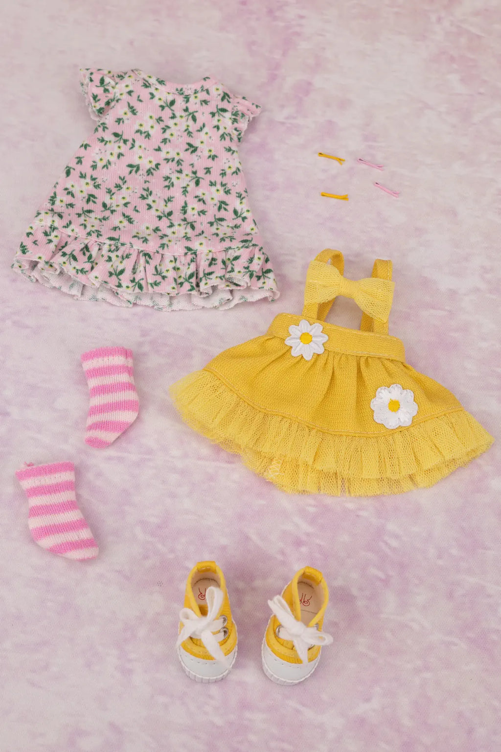 A doll clothing set laid out on a pink textured background, including a pink floral dress, a yellow pinafore-style dress, pink striped socks, and yellow sneakers with white laces.