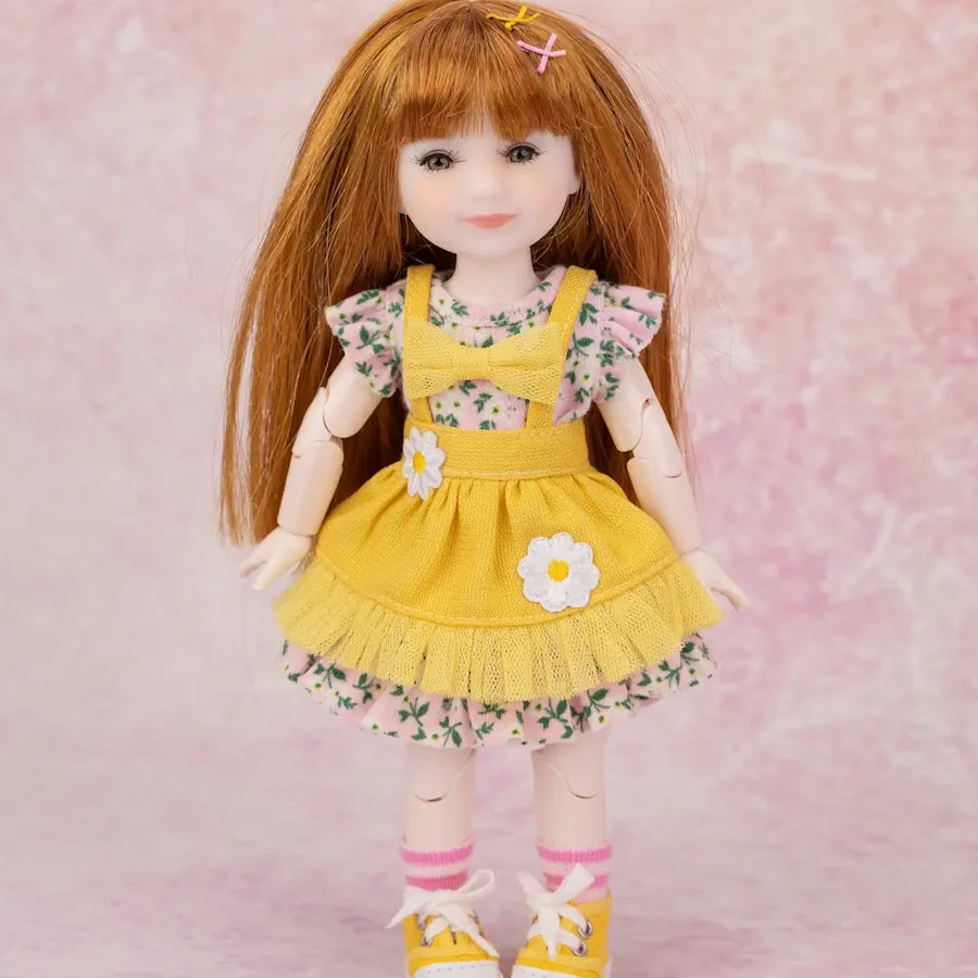 A doll with long auburn hair wearing a yellow floral dress with white ruffled trim, pink floral undershirt, pink socks, and yellow shoes, posed against a soft pink textured background.