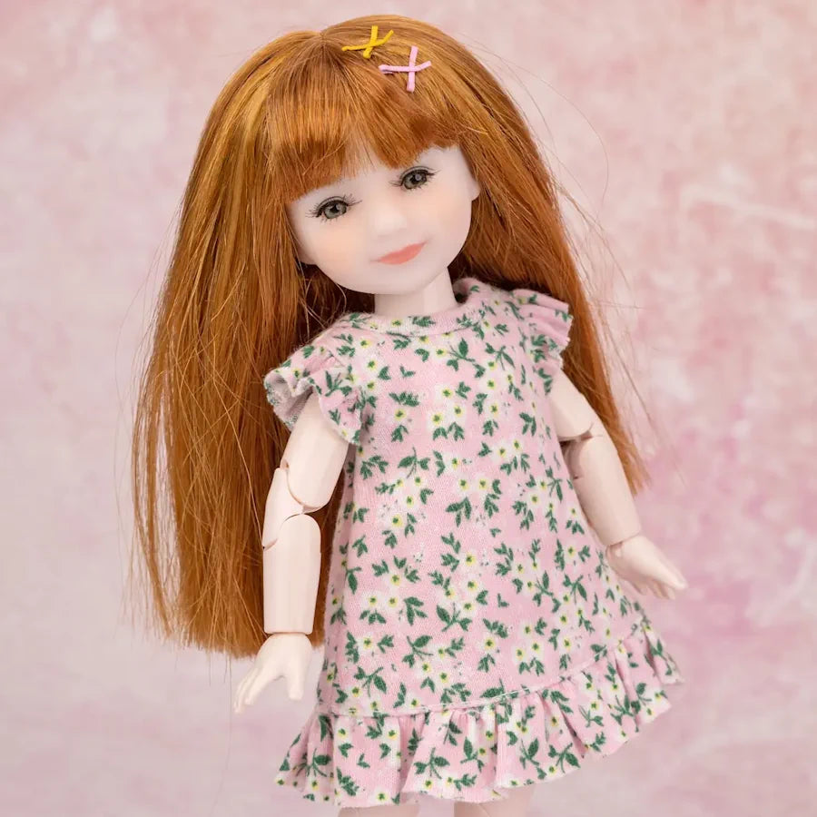 A doll with long auburn hair wearing a pink dress with a green and white floral print, posed against a soft pink textured background.