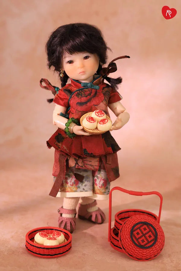 A doll dressed in a red traditional outfit stands against a warm beige backdrop, holding two small round pastries. A matching red woven basket and a small red tray with another pastry are placed in front of it.