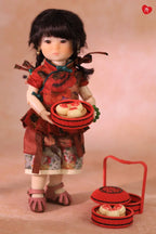 A doll dressed in a red traditional outfit holding a red basket of pastries, with a matching red basket and lid placed beside it on a soft beige studio background.