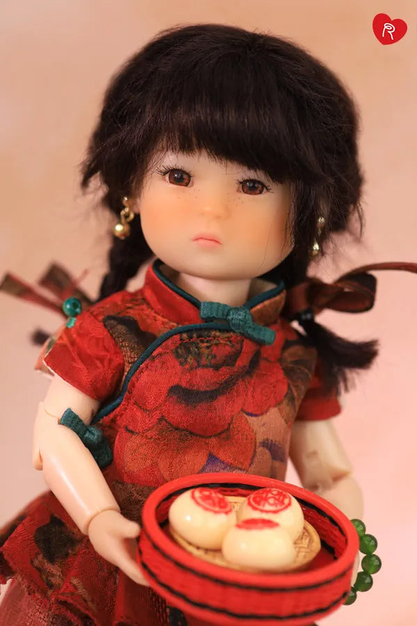 A doll with dark hair wearing a red floral-patterned dress, holding a small red tray with dumplings against a soft pink background.