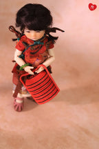 A doll with dark hair wearing a red floral cheongsam dress and holding a red striped basket, posed against a soft beige background.