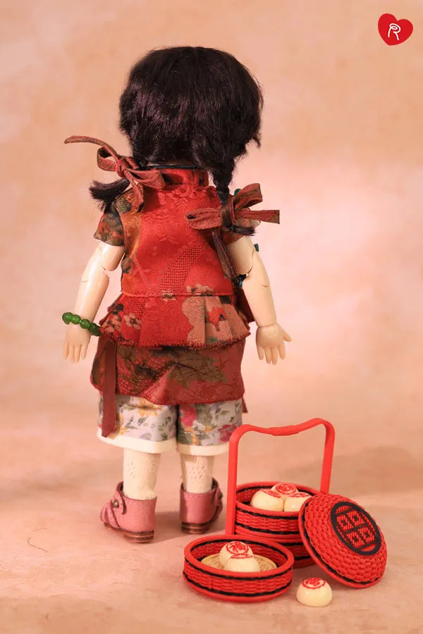 A doll viewed from behind wearing a red patterned traditional-style top and floral shorts, standing on a peach studio background with a small red basket and miniature accessories beside it.