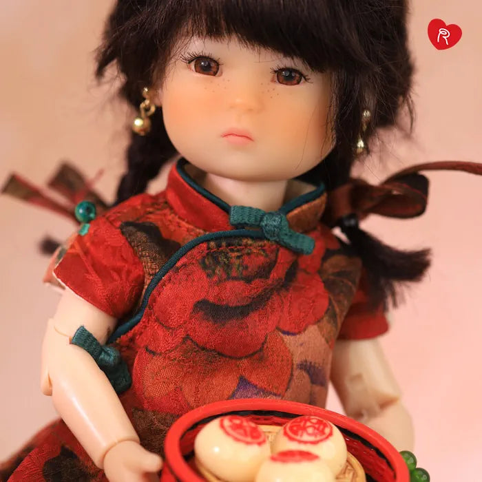 A close-up of a Ten Ping doll with dark hair wearing a red floral cheongsam-style dress and holding a red tray with round pastries, against a soft pink background.