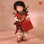 A doll with black hair stands holding a red striped basket against a warm beige studio background. The doll wears a red floral dress, patterned skirt, white socks, and pink shoes.