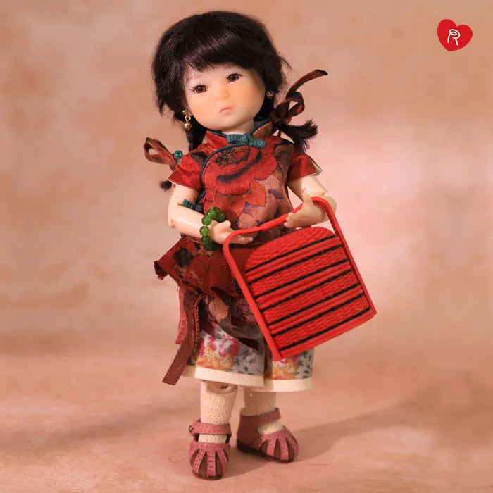 A doll with black hair stands holding a red striped basket against a warm beige studio background. The doll wears a red floral dress, patterned skirt, white socks, and pink shoes.